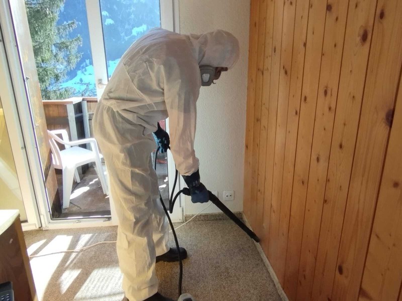 Dans un appartement a Lausanne, un deratiseur en combinaison de protection applique un traitement contre les nuisibles le long d’un mur en bois pres de la porte-fenetre.